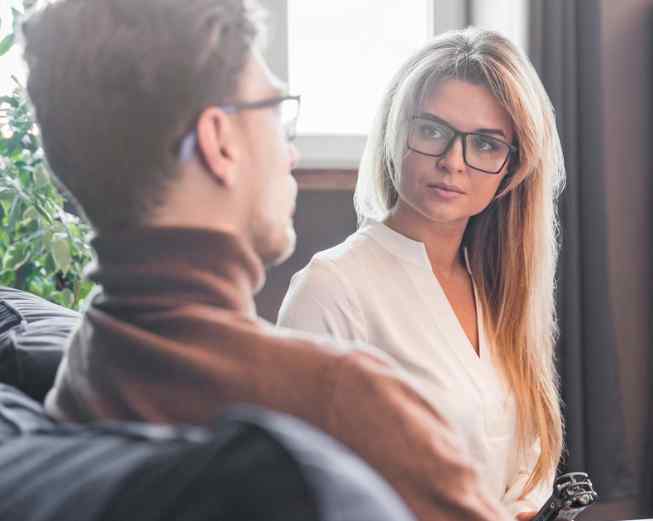 Two people wearing glasses sit indoors, facing each other and having a conversation. The woman listens attentively to the man.